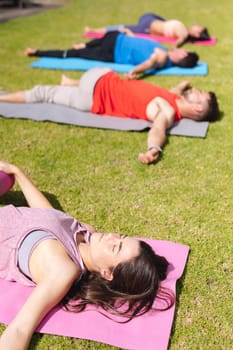 Young woman lying on exercise mat with eyes closed while practicing yoga with people in park. healthy lifestyle and body care.