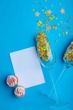 Champagne flutes with confetti by cupcakes and blank paper with copy space on blue background. party, celebration and desert.