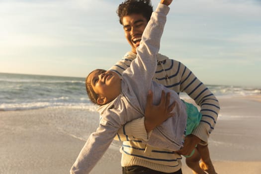 Cheerful biracial man carrying daughter pretending to fly at beach against sky on sunny day. family, lifestyle and weekend.