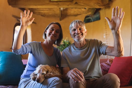 Portrait of mature couple with dog waving hands while sitting on couch in cob home. togetherness, lifestyle and sustainable living.