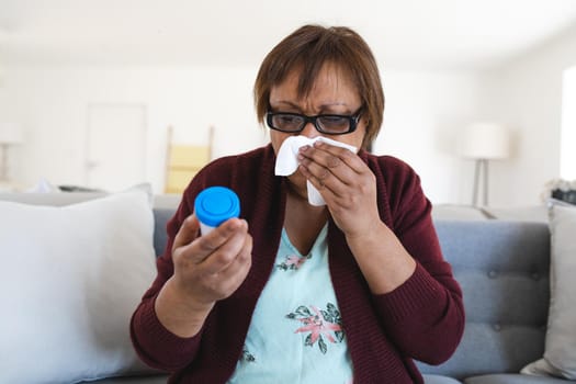 African american senior woman suffering with cold and flu holding medicine container at home. lifestyle, illness and healthcare.