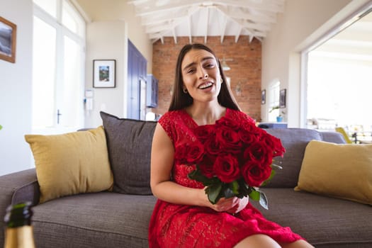 Portrait of smiling beautiful biracial woman holding rose bouquet during virtual date at home. lifestyle and virtuality.