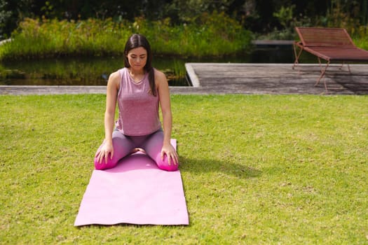 Young woman kneeling on exercise mat while practicing yoga in park on sunny day. healthy lifestyle and body care.