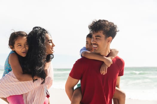 Happy biracial father and mother giving piggyback rides to children at beach against sky. family, lifestyle and weekend.
