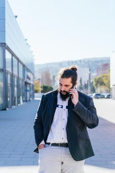 Young businessman with long hair and beard calling during his work break, on the street next to the offices. Vertical photo on a sunny and clear day. Front view