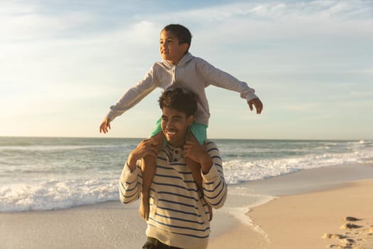 Happy biracial father carrying son on shoulders with arms outstretched enjoying sunset at beach. family, lifestyle and weekend.