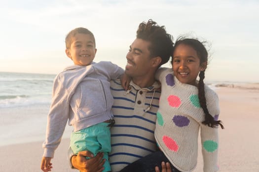 Happy biracial father carrying children while standing at beach against sky during sunset. family, lifestyle and weekend.