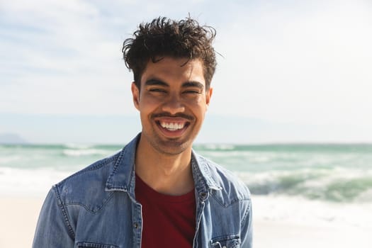 Portrait of smiling biracial man wearing denim shirt at beach on sunny day. lifestyle and weekend.