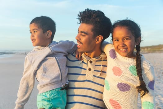 Happy biracial man carrying children while standing at beach against sky during sunset. family, lifestyle and weekend.