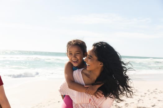Happy biracial woman giving piggyback ride to daughter at beach against sky with copy space. family, lifestyle and weekend.
