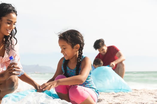 Happy biracial mother and daughter collecting waste plastic bottle in bag at beach on sunny day. lifestyle and environmentalism.