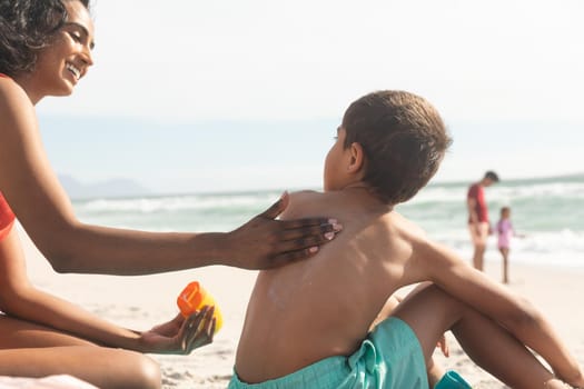 Smiling biracial woman applying sunscreen lotion on shirtless son's back at beach during sunny day. family, lifestyle and care.
