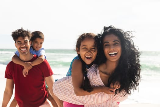 Portrait of happy multiracial parents giving piggyback rides to kids at beach with copy space. family, lifestyle and weekend.