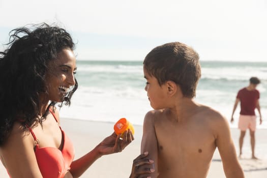 Shirtless biracial boy looking at mother with sunscreen lotion bottle on sunny day. family, lifestyle and care.