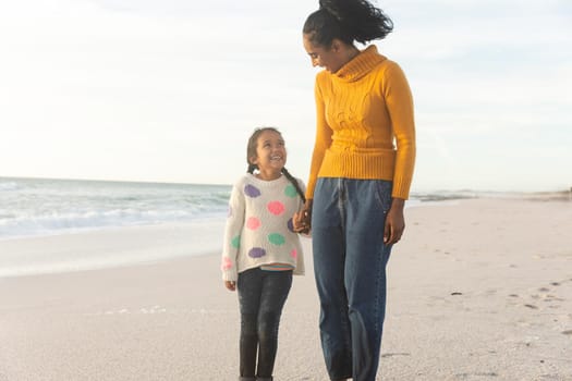 Happy biracial mother and daughter holding hands while looking at each other on beach against sky. family, lifestyle and weekend.