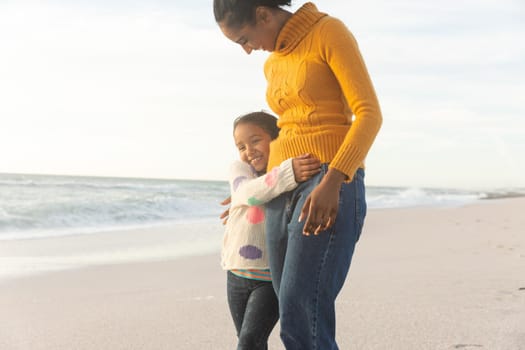 Happy biracial girl embracing mother while walking at beach against sky during sunset. family, lifestyle and weekend.