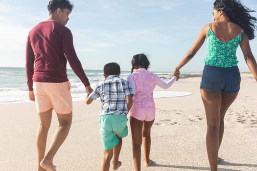 Rear view of biracial parents holding hands with children walking on sand at beach during sunny day. family, lifestyle and weekend.