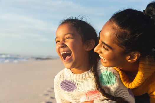 Cheerful young biracial woman with daughter laughing at beach against sky during sunset. family, lifestyle and weekend.