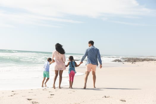 Full length rear view of multiracial family holding hands walking on shore at beach during sunny day. lifestyle and weekend.