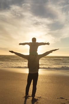 Rear view of biracial man carrying son on shoulders with arms outstretched at beach during sunset. family, lifestyle and weekend.