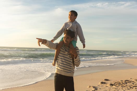 Happy biracial father pointing to son while carrying him on shoulders at beach against sky. family, lifestyle and weekend.