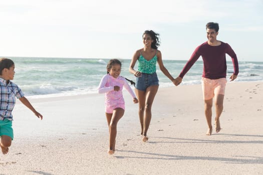 Biracial children running ahead of smiling parents on shore at beach against sky during sunny day. family, lifestyle and weekend.