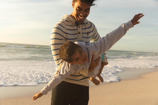 Happy biracial man carrying son pretending to fly with arms outstretched at beach during sunset. family, lifestyle and weekend.