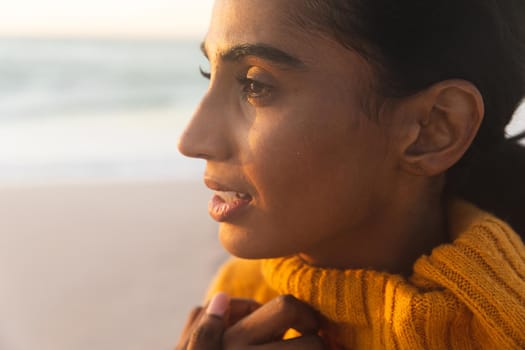 Close-up of young thoughtful biracial woman in sweater looking away at beach during sunset. lifestyle and weekend.