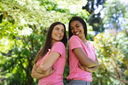 Happy multiracial female friends with arms crossed wearing breast cancer awareness ribbons in park. breast cancer awareness campaign, friendship and unity concept.