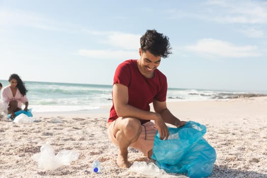 Smiling biracial man collecting garbage in blue plastic bag from sand at beach against sky. lifestyle and environmentalism.