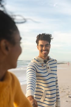 Smiling biracial man holding hand of girlfriend while standing behind her at beach against sky. lifestyle, love and weekend.