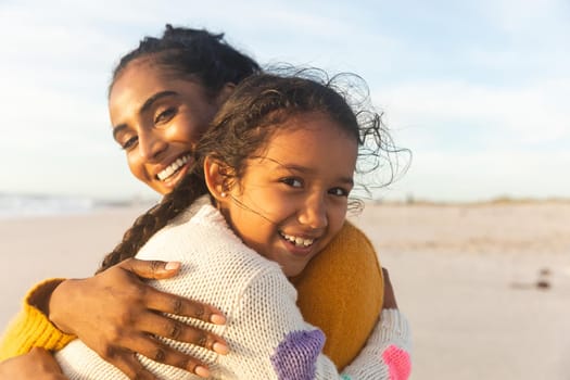 Portrait of smiling biracial mother and daughter embracing each other at beach during sunset. family, lifestyle and weekend.