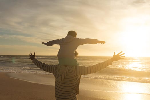 Rear view of biracial man carrying son on shoulders with arms outstretched at beach during sunset. family, lifestyle and weekend.