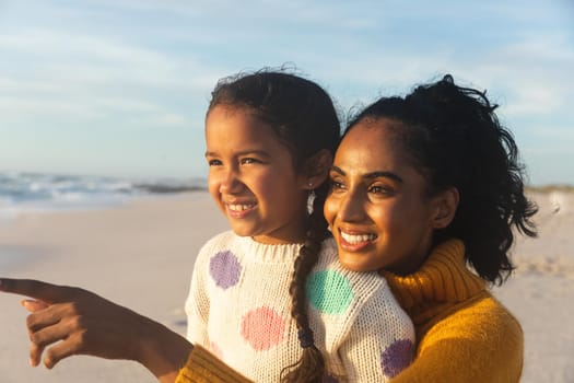 Happy biracial woman pointing to daughter at beach against sky during sunset. family, lifestyle and weekend.