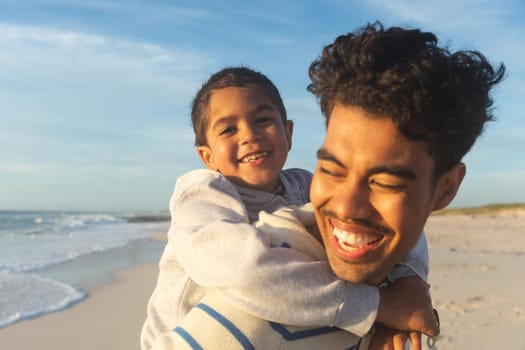 Close-up of happy biracial father giving son piggyback ride to son at beach against sky. family, lifestyle and weekend.