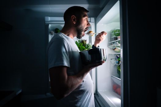 Hungry man eating food at night from open fridge. Man taking midnight snack from refrigerator. High quality photo