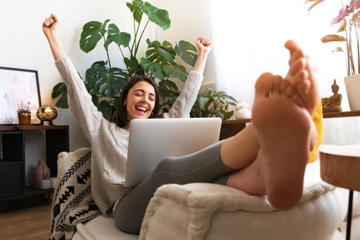 Excited and happy young caucasian woman raising arms up celebrating success or achievement at home using laptop. Victory concept.