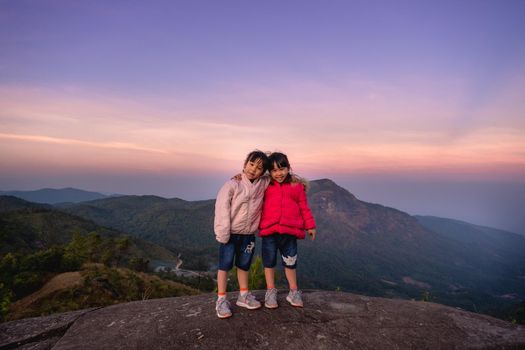 Two cute little girls hugging on top of a mountain and looking at the camera against a sunset background. Hiking adventures with family on vacation.
