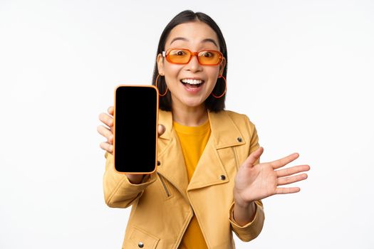 Enthusiastic asian female model, showing smartphone app interface, online store or website on mobile phone screen, standing over white background.