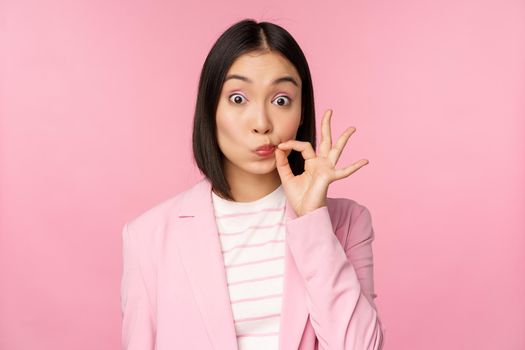 Portrait of asian corporate woman showing mouth seal, close shut lips on key gesture, promise keep secret, standing over pink background in suit.