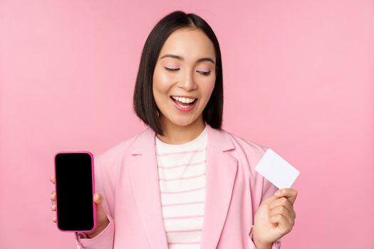 Smiling korean businesswoman in suit, showing mobile phone screen, credit card, showing online banking application interface, pink background.