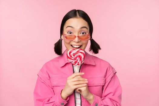 Silly and cute asian female model licking lolipop, eating candy sweet and smiling, looking excited, standing over pink background.