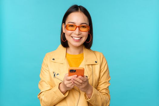 Smiling asian girl in sunglasses, using smartphone app, holding mobile phone, standing over blue background. Copy space