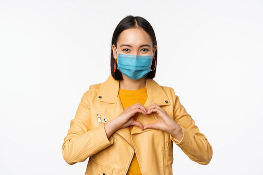 Covid-19 and people concept. Young asian woman in medical face mask, showing heart gesture and smiling with care, white background.