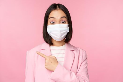 Portrait of asian businesswoman in medical face mask and suit, pointing finger left, showing advertisement, company banner, studio pink background.