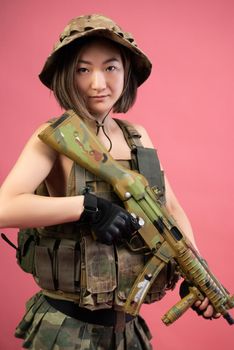 the sexy Asian woman in military clothes with an automatic rifle in her hands on a pink background