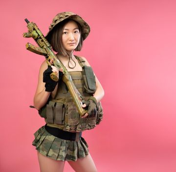 the sexy Asian woman in military clothes with an automatic rifle in her hands on a pink background