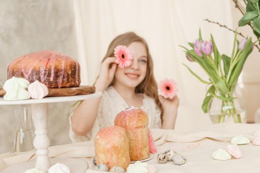 A girl with long hair in a light dress is sitting at the Easter table with cakes, spring flowers and quail eggs. Happy Easter celebration
