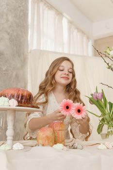 A girl with long hair in a light dress is sitting at the Easter table with cakes, spring flowers and quail eggs. Happy Easter celebration
