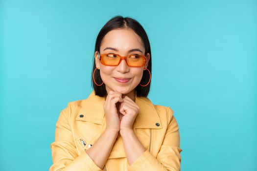 Portrait of stylish korean woman in sunglasses, smiling and looking aside at logo coquettish, standing over blue background. Copy space